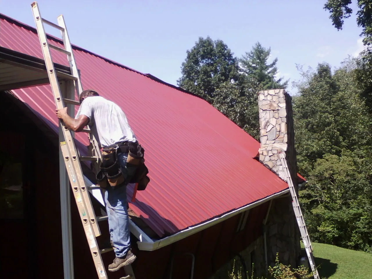 Roofer on a ladder beside a red metal roof and stone chimney — Soffit & Fascia Repair by Roofing Brothers in Chadbourn